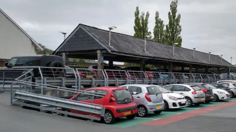 Large outdoor car park on a cloudy day, featuring a long covered parking structure (with a slanted roof) with multiple vehicles parked underneath and in marked bays beside it. A black SUV is parked on an elevated ramp in the foreground, while rows of cars line the car park. A residential building with a white wall and tall trees are visible in the background under an overcast sky.