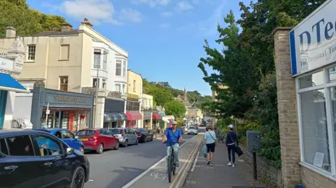 John Wimperis A view of Hill Road in Clevedon. A cyclist is coming towards the camera and a black car is moving away. There are shops on either side of the road.