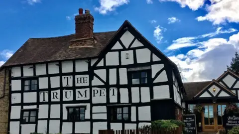 The Trumpet Inn A tudor framed black and white pub, painted with a title saying The Trumpet on the front. The building has five windows at the front and a side entrance to its left. There is a wavy tiled roof with a large chimney and a blue sky back drop.