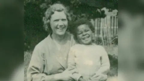 Fumi Stephenson A black and white photo of Dr Stephenson as a young child, wearing a light coloured jumper. He is being held by his foster mother in a garden. Both are laughing and smiling at the camera. 