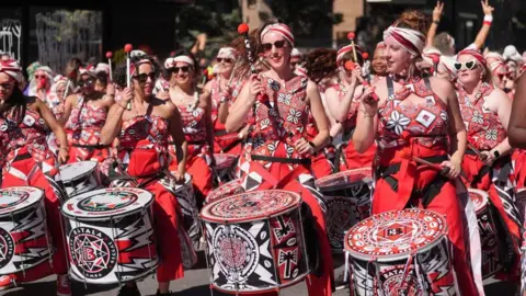 PA Media Members of a drumming troupe perform during a carnival parade, wearing coordinated red, black and white costumes and headscarves while playing decorated percussion drums in a city street.