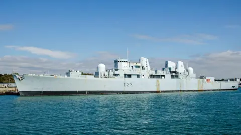 An enormous grey warship docked on a quayside on a sunny day. The hull has a couple of rust marks.
