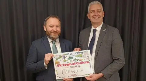 David Smith David Smith at Parliament. He has short grey hair and is smiling. He and another man, on his left, are both wearing suits and are holding a sign stating they are backing Berwick for UK Town of Culture.