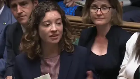 A woman with mid-length curly brown hair, speaking in parliament. A male and female MP can be seen behind her. Coombes is wearing a navy suit jacket and pink striped top.