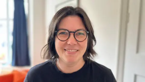 Molly Haworth, head and shoulders shot, showing a lady with dark hair and glasses smiling at the camera in a black t-shirt