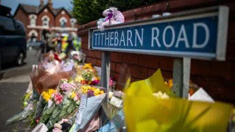 A row of flowers lay next to a small brick wall and a sign reading 'Tithebarn Road'