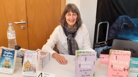 SUSAN PAPE Author and journalist Susan Pape - pictured wearing a white blouse and scarf - sits behind a table with some of her books.