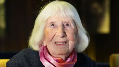 PA Media Betty Brown has shoulder-length light-colored hair and is sitting on a yellow cushioned chair. She is wearing a dark textured jacket, a pink and purple silk scarf around the neck. The background is dark and blurred.