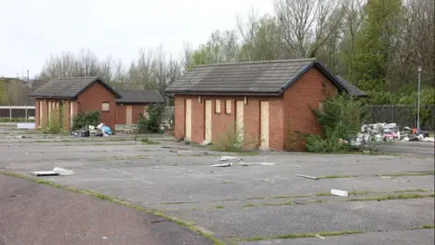 LDRS Two abandoned brick buildings, with bordered up windows. Rubbish sits behind them.