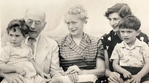 Moritz Family Photo A black and white image of the Moritz family. Sir Michael is a child in this image and is pictured on the far right sitting on his mothers knee, he has dark hair and a white patterned shirt on, as well as dark shorts. His mother looks to the left of the image and is wearing a black and white patterned dress. She too has dark hair. In the middle is Sir Michael's grandmother, who has a black and white striped dress, her hands are atop a white purse and she has short, white, curly hair. Next to her is a man, Sir Michael's grandfather, who is bald and has round-framed glasses on. He has a white suit jacket, over a white shirt and a lightly coloured tie. He has a baby girl on his lap who has a light-coloured dress on and dark curly hair.