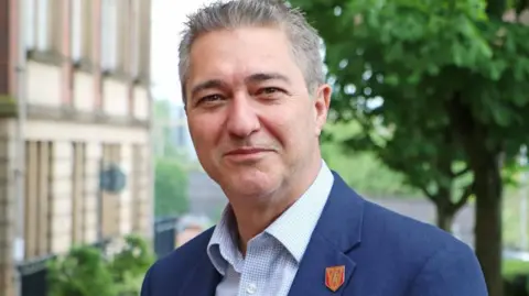 LCC Head and shoulders image of Mark Wynn. He is smiling, has short grey hair and is wearing a blue suit and pale blue checked shirt