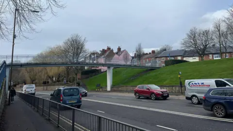 LDRS A grey bridge with blue railings suspended over a busy road with traffic moving in both directions. A grassy bank and houses can be seen to the right.