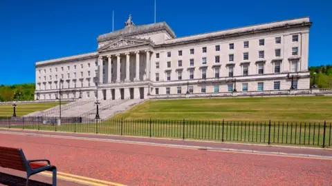 A large white/grey building it has green grass outside it and a red road