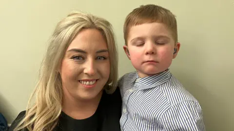 Gemma is holding her son George in the picture. She has long blonde hair and is wearing makeup and a black t-shirt. George has light brown hair and is wearing a blue and white striped shirt. 