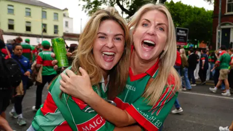 PA Media Two people stand in the street, excitement on their faces. They both have blonde hair and are wearing green and red jerseys. Behind them there is a crowd of people walking, many of whom are also wearing green and red jerseys. 