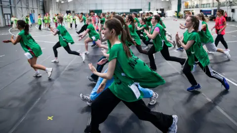 Glasgow 2026 A group of dancers, mostly wearing green bibs, practice a routine inside a sports hall