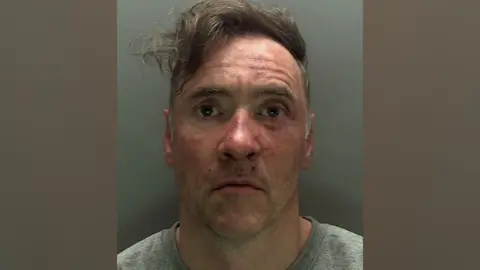 CPS Uma foto policial de Paul Doyle vestindo um suéter cinza e parecendo perplexo. Seu cabelo, com franja longa e castanha, está bagunçado e de lado. Existem manchas vermelhas em seu rosto