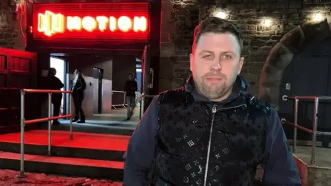 Daniel Deeks stands in front of the Motion nightclub at night. He is wearing dark clothing and looking at the camera.