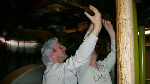 Dan Button Two people dressed in white overalls and hair nets look at a pipe.