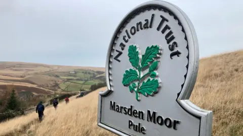 A cast-iron sign with the words Marsden Moor and Pule on the bottom and National Trust running around the top. There are some walkers in dark clothing in the background walking along a moorland path. 