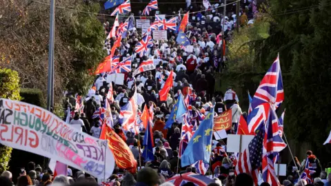 Reuters A large group of protestors. They are holding Union Jack and local flags. 