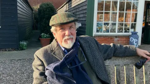 Vikki Irwin/BBC Reggie Oliver wears a tweed coat, a blue scarf and a tweed flat cap. He sits in front of a shop on a bench. He has glasses around his neck and a white beard