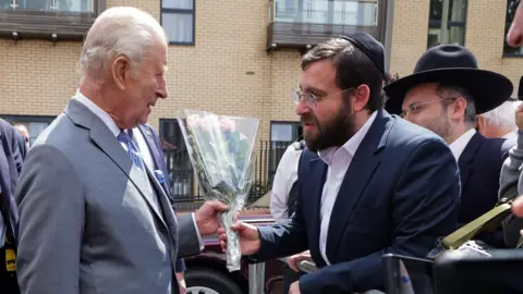 Getty Images King Charles III in a grey suit is presented with flowers by a Jewish member of the community of Golders Green who is standing behind a barrier with a crowd of other Jewish people.