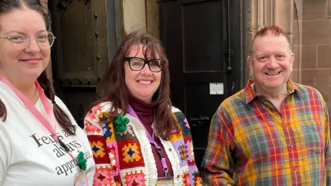 Jonny Humphries/BBC Two women, one with brown hair in pigtails and one with long dark brown hair, and a man with hair dyed red, smile at the camera outside the front door of a church. 