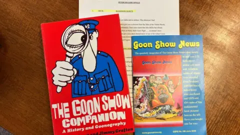 BBC/Fiona Callow Three items of The Goon Show memorabilia is displayed on a wooden table. On the left, a red book with a cartoon of a policeman looking through a magnifying glass titled 'The Goon Show Companion'; in the centre and covered by the other two items, the script for a sketch, and on the right a blue magazine-style newsletter titled 'Goon Show News'.