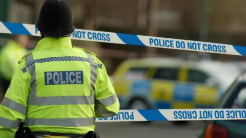 BBC A police officer wearing a yellow jacket standing in front of a police cordon 