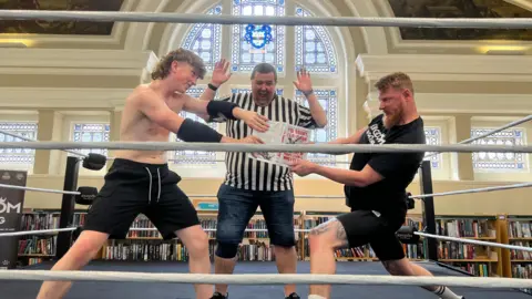 A boy in black shorts and no shirt stands on the left, a man wearing black shorts and a black T-shirt is on the right. They are both holding onto a book, fighting over it. Behind them stands a ref. All three are standing in a boxing ring. Behind them is a large, arched stained glass window above bookshelves.