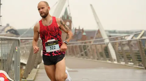 Derry City and Strabane District Council Kyle running over a bridge. He is wearing a red top and black shorts.