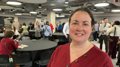 Amy Holmes/BBC A woman wearing a burgundy top stands in the foreground of a busy event space. Behind them, groups of people are gathered around round tables covered with black tablecloths. Attendees are chatting, checking phones, and moving around the large room, which has exposed ceilings, circular light fixtures, and a modern industrial design.