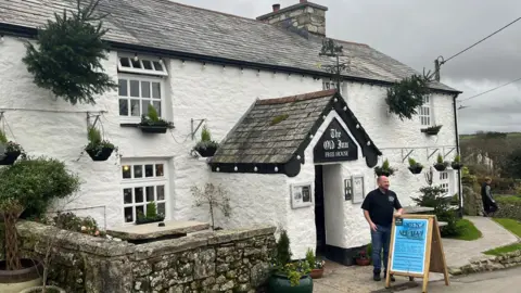 A pretty white pub with black guttering and porch and a slate roof, there are Christmas trees in pots on the walls and a man is standing in the doorway by a blue advertising sign 