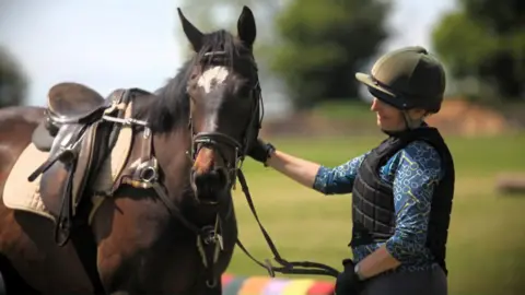 Sue Ravenhill-Handley Sue is wearing horse riding gear and petting her horse, who is brown and has a white patch on its face.