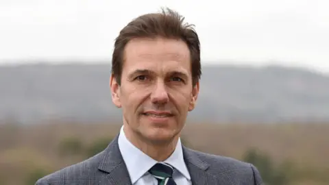 Simon Bowden Simon Bowden a man in a grey suit with a white shirt and black, green and white tie.