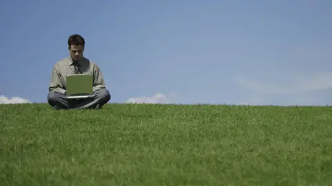 PA Media A young man is sitting cross-legged on the brow of a grassy hill typing on a laptop. He is wearing trousers, a shirt and a tie and his head is bowed as he types. It is a clear blue sky with a couple of wispy clouds.