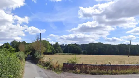 Google A narrow country road with large field to the right with trees and bushes surrounding on either side.