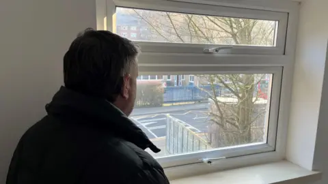 Brett Kemp, a man with short grey hair wearing a dark blue jacket, standing in an upstairs room inside his house, looking over the toilet and a school on the opposite side of the road