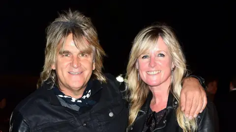 PA Media Mike Peters and his wife Jules attending the premiere of Soul Boys of the Western World, Spandau Ballet. They are both wearing leather jackets, with a black background behind them. Mike has his trademark shoulder length hair, while Jules has long blonde hair reaching past her shoulders and a side fringe.