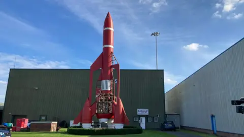 A 60ft tall red, Thunderbird rocket. It's on a grass field in front of a green building. It has white accent stripes