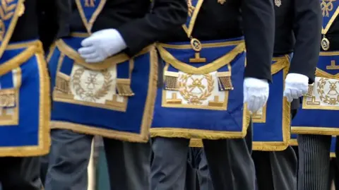 PA Media Masons march in a procession. Pictured are men from waist height wearing white gloves and wearing the Freemasons coat of arms embraided with gold thread on a royal blue background
