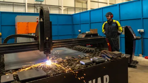A man in high vis protection gear, including a cap and ear defenders, stood in front of a factory line where metal is being shapes by a machine and sparks are flying off it.