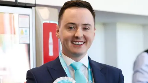 Reform deputy leader Darren Grimes. He has short brown hair that are shorter on the sides and combed to one side. He has green eyes and is smiling. He wears a navy suit, with light blue tie, lanyard and rosette. The background is blurred.