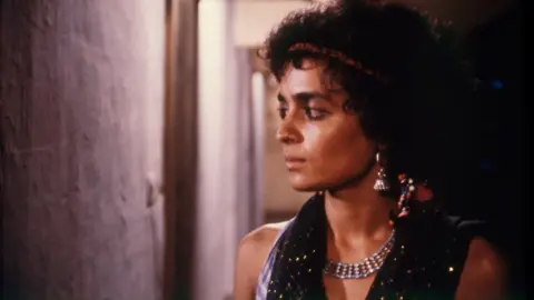 Film Heritage Foundation A still of Arundhati Roy, wearing silver danglers and a necklace and a black tank top, as she looks sideways 