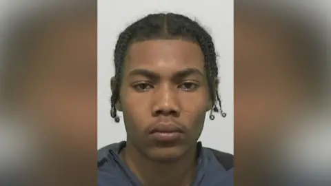 Mugshot of Mohammed Moufareh. He has a very faint moustache and black hair in plaits.