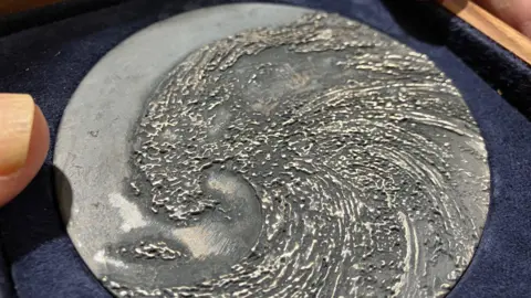 Jon Wright/BBC Silver-coloured metal disc with a raised image of a wave engraved on it