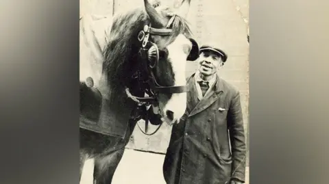 Benson family photograph Black and white still of carter Dick Benson dressed in a coat, scarf and cap smiling as he stands next to a horse holding the reins.