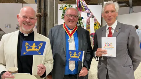 Jon Wright/BBC Three men stand with some art displayed in the background. The man on the left has a black shirt and white dog collar; centre is a man in a blue shirt and grey suit jacket; on the right is a man with white hair and a grey suit jacket.