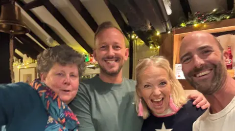 A lady with short curly hair wearing a blue and red scarf (pub volunteer Geraldine Symonds) pictured smiling with three other local members of the community - two men and a woman. They are standing in the Swan Pub with Christmas fairy lights and the pub bell in the background. 